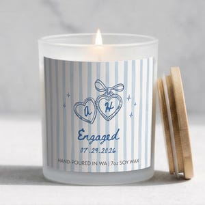 May include: A frosted glass candle with a light blue and white striped label featuring a blue heart locket illustration, the letters A and H, the text Engaged, the date 07.29.2026, and the text Hand-poured in WA | 7oz soy wax, with a wooden lid.