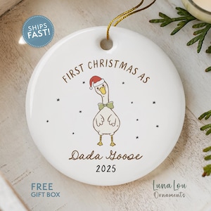 May include: White ceramic Christmas ornament with a cartoon goose wearing a Santa hat and bow tie. Text reads "First Christmas as Dada Goose 2025". Includes a gold string for hanging. The ornament is a holiday decoration.