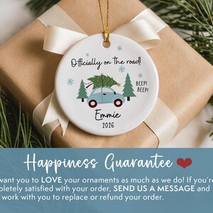 May include: A round white ceramic ornament featuring a light blue car carrying a green Christmas tree on its roof. The text reads Officially on the road!, Emmie, and 2026, accompanied by small blue snowflakes and green pine trees. It hangs by a gold cord.