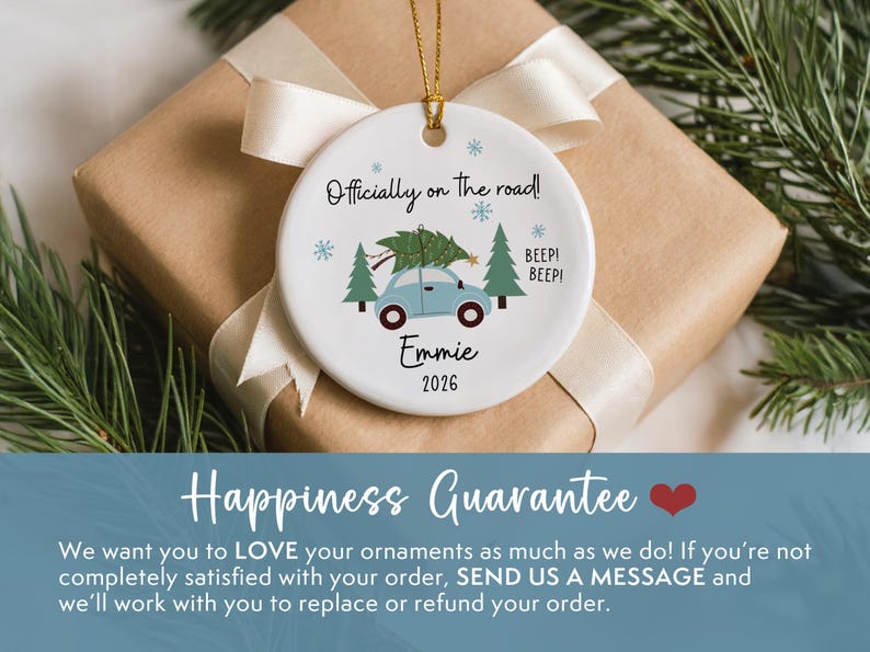 May include: A round white ceramic ornament featuring a light blue car carrying a green Christmas tree on its roof. The text reads Officially on the road!, Emmie, and 2026, accompanied by small blue snowflakes and green pine trees. It hangs by a gold cord.