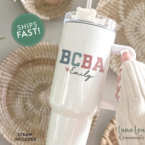 May include: White tumbler with a handle, featuring the text "BCBA" in blue and red, with a small heart and the name "Emily" in script. The tumbler includes a straw and is held by a person wearing a white sweater.