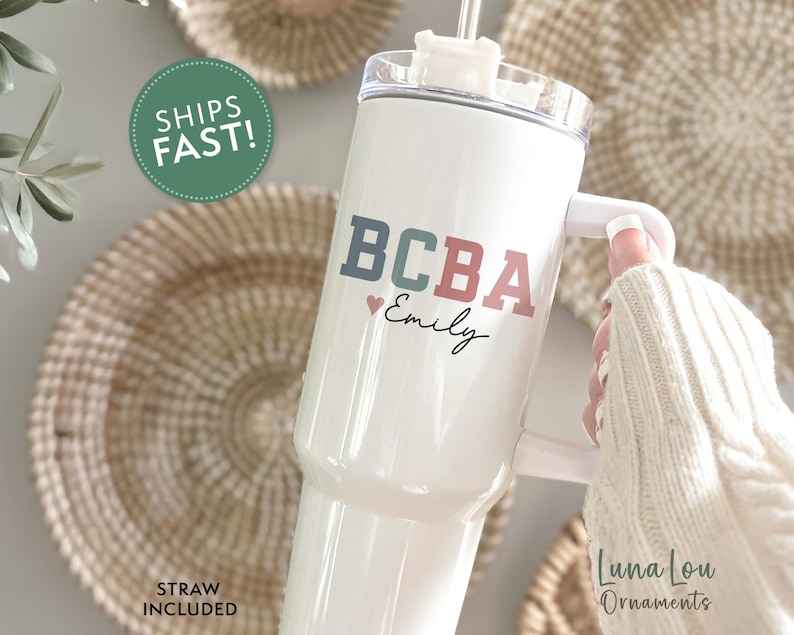 May include: White tumbler with a handle, featuring the text "BCBA" in blue and red, with a small heart and the name "Emily" in script. The tumbler includes a straw and is held by a person wearing a white sweater.