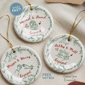 May include: Three round white ceramic ornaments with gold hanging strings. Each features a green wreath design, red text with names, the word Engaged, and a date. Graphics include a ring box, hands with a ring, and champagne flutes.