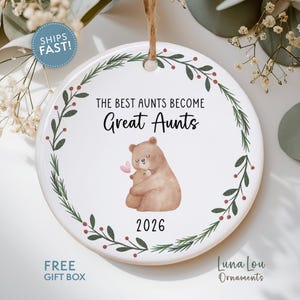 May include: A round white ceramic ornament featuring a watercolor illustration of two brown bears hugging with a small pink heart. The text reads THE BEST AUNTS BECOME Great Aunts and 2026. A green pine branch wreath with red berries encircles the design.