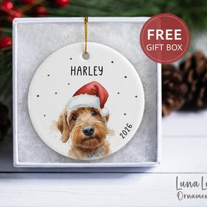 May include: A round white ceramic ornament featuring a watercolor illustration of a golden-brown dog wearing a red and white Santa hat. The name HARLEY is printed above the dog, and the year 2026 is printed to the right. A gold string is attached.