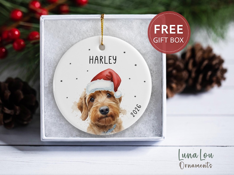 May include: A round white ceramic ornament featuring a watercolor illustration of a golden-brown dog wearing a red and white Santa hat. The name HARLEY is printed above the dog, and the year 2026 is printed to the right. A gold string is attached.