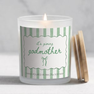May include: A lit soy wax candle in a frosted glass jar with a wooden lid. The label features the text "it's giving godmother" in green script, set against a white background with green stripes. The candle is 198g.