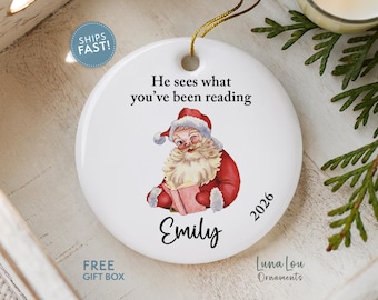May include: A round white ceramic ornament featuring a vintage-style illustration of Santa Claus reading a book. The text reads, He sees what you’ve been reading, with the name Emily and the year 2026 printed in black script and sans-serif fonts.