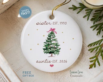 May include: A round white ceramic ornament featuring a watercolor green Christmas tree topped with a pink bow. The text reads "sister EST. 1993" above the tree and "auntie EST. 2026" below it, accompanied by a small pink heart and gold star accents.