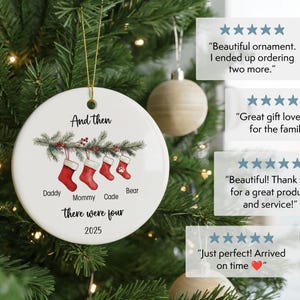 May include: A white ceramic Christmas ornament with a gold string, hanging on a Christmas tree. The ornament features red stockings, text that reads "And then there were four 2025", and names: Daddy, Mommy, Cade, and Bear.