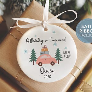 May include: A round white ceramic ornament featuring a pink car carrying stacked gifts between two green pine trees. The text reads Officially on the road!, BEEP! BEEP!, Olivia, and 2026, accented by blue snowflakes and a white satin ribbon.