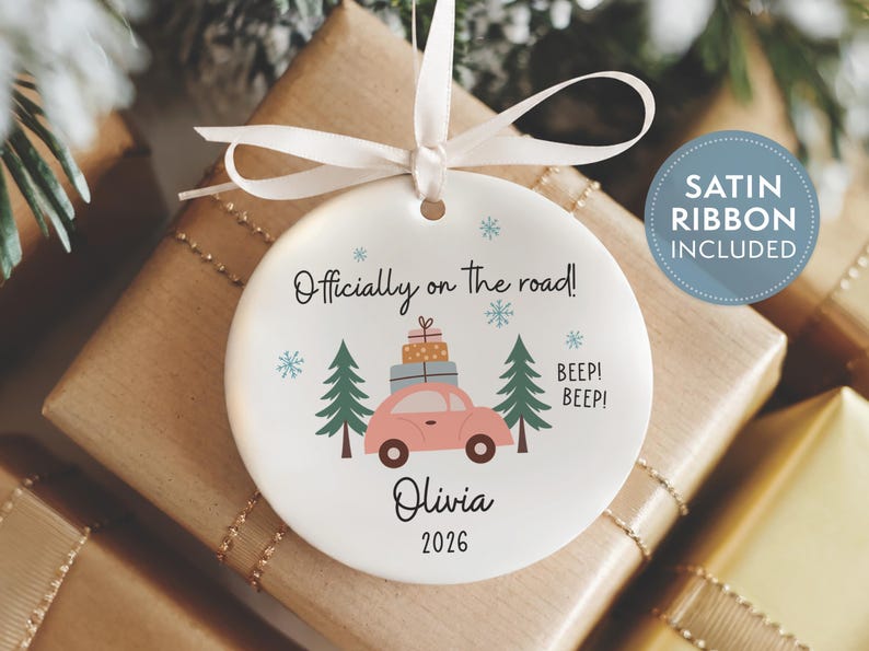 May include: A round white ceramic ornament featuring a pink car carrying stacked gifts between two green pine trees. The text reads Officially on the road!, BEEP! BEEP!, Olivia, and 2026, accented by blue snowflakes and a white satin ribbon.