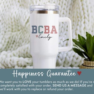 May include: White tumbler with a handle, featuring the text "BCBA" in blue, green, and red, with "Emily" below. A small heart symbol is included. The tumbler has a clear lid and is set against a white knitted background.