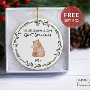 May include: A white ceramic ornament with a gold string, featuring a bear hugging a baby bear, a floral wreath, and the text "The Best Grandmas Become Great Grandmas 2025." The ornament is in a gift box with a "FREE GIFT BOX" sticker.