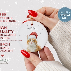 May include: A round white 3 inch ceramic ornament featuring a printed illustration of an orange tabby cat wearing a red and white Santa hat. The name MITTENS is printed in black above the cat, surrounded by small black dots on the surface.
