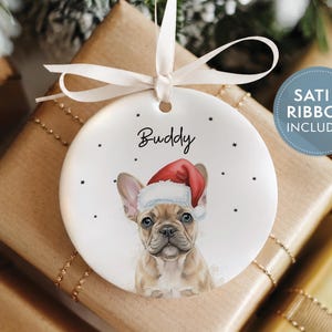 May include: A round white ceramic ornament featuring a tan French Bulldog wearing a red and white Santa hat. The name Buddy is printed in black above the dog. The ornament is suspended by a white satin ribbon and includes small black star accents.