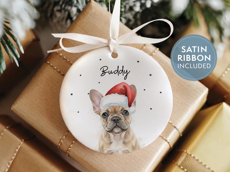 May include: A round white ceramic ornament featuring a tan French Bulldog wearing a red and white Santa hat. The name Buddy is printed in black above the dog. The ornament is suspended by a white satin ribbon and includes small black star accents.