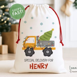 May include: A white Christmas gift bag with a red drawstring features a yellow truck carrying a green Christmas tree. The bag is decorated with snowflakes and the text "SPECIAL DELIVERY FOR HENRY".