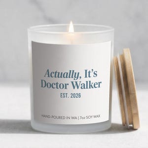 May include: A lit, frosted glass candle with a wooden lid. The candle has a white label with the text "Actually, It's Doctor Walker EST. 2026". The candle is hand-poured in WA and contains 7oz of soy wax.