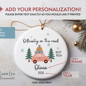 May include: A round white ceramic ornament featuring a pink car illustration with stacked gifts on its roof, flanked by two green pine trees. The text reads "Officially on the road!", "BEEP! BEEP!", "Olivia", and "2026". It includes a gold hanging string.
