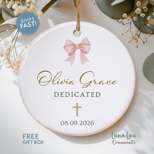 May include: A round white ceramic ornament featuring a pink bow graphic at the top. The text reads Olivia Grace in gold script, followed by DEDICATED in gray, a gold cross symbol, and the date 08.09.2026 in black. It hangs from a brown twine loop.