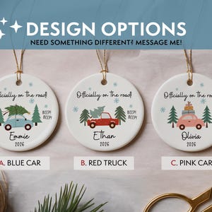 May include: Three round white ceramic ornaments with gold hanging strings. Each features the text 'Officially on the road!' and 'BEEP! BEEP!'. Designs include a blue car, a red truck, and a pink car, each with trees, snowflakes, names, and 2026.