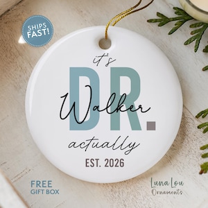 May include: A round white ceramic ornament featuring the text "it's DR. Walker actually" in a mix of blue, black, and grey fonts. The design includes the year "EST. 2026" in grey at the bottom and a gold hanging string at the top center.