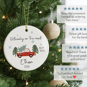 May include: A round white ceramic ornament featuring a red truck carrying a Christmas tree, with the text "Officially on the road!", "Ethan", "2026", and "BEEP! BEEP!" along with blue snowflakes and green pine trees, hanging by a gold string.