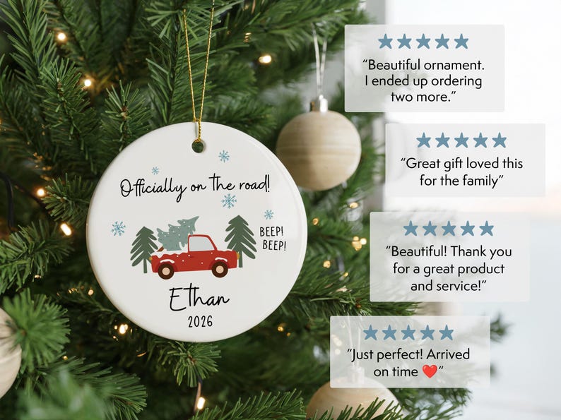 May include: A round white ceramic ornament featuring a red truck carrying a Christmas tree, with the text "Officially on the road!", "Ethan", "2026", and "BEEP! BEEP!" along with blue snowflakes and green pine trees, hanging by a gold string.