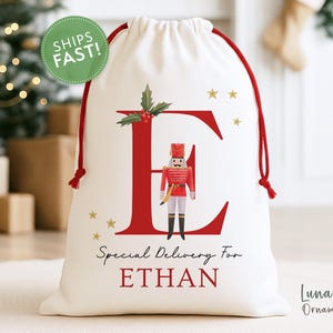 May include: A white drawstring gift bag with a red letter "E" and a toy soldier design. The bag has a red drawstring and the text "Special Delivery For ETHAN". Gold stars and holly accents decorate the bag.