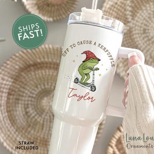May include: White tumbler with a handle and a straw. The tumbler features a cartoon frog riding a scooter, wearing a red hat, and the text "OFF TO CAUSE A KERFUFFLE" and "Taylor". The tumbler has a white handle.