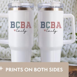 May include: Two white travel mugs with handles, each featuring the text "BCBA" in blue, green, and pink, with "Emily" below. A small pink heart is between the text and the name. The mugs have clear lids and are on a white background.