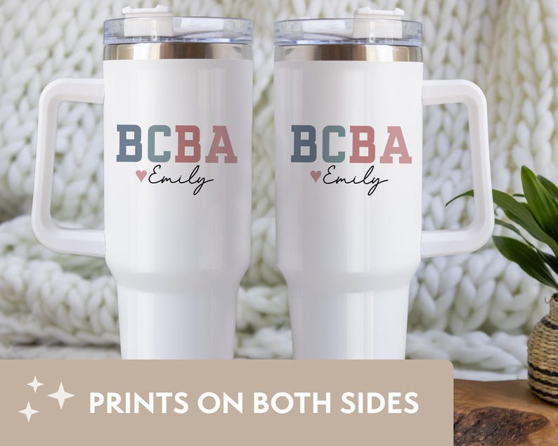May include: Two white travel mugs with handles, each featuring the text "BCBA" in blue, green, and pink, with "Emily" below. A small pink heart is between the text and the name. The mugs have clear lids and are on a white background.