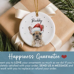 May include: A round white ceramic ornament featuring a tan French Bulldog wearing a red and white Santa hat. The name Buddy is printed in black script above the dog, surrounded by small black dots. A gold string is attached for hanging.