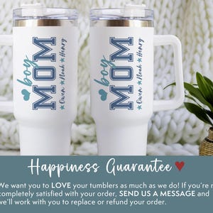 May include: Two white travel mugs with black handles. The mugs have a blue and white design that says "boy MOM" with a star and the name "Owen Noah Henry" underneath. The mugs are sitting on a white fuzzy surface.