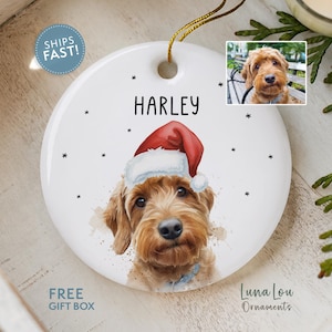May include: A round white ceramic ornament featuring a watercolor-style illustration of a golden-brown dog wearing a red Santa hat. The name HARLEY is printed in black above the dog. A small rectangular photo of the dog is inset at the top right.