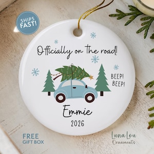 May include: A round white ceramic ornament featuring a light blue car carrying a green Christmas tree on its roof. The text reads Officially on the road!, BEEP! BEEP!, Emmie, and 2026. Blue snowflakes and green pine trees decorate the design.