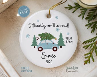 May include: A round white ceramic ornament featuring a light blue car carrying a green Christmas tree on its roof. The text reads Officially on the road!, BEEP! BEEP!, Emmie, and 2026. Blue snowflakes and green pine trees decorate the design.