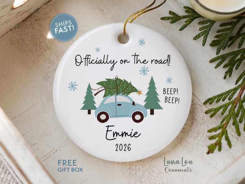 May include: A round white ceramic ornament featuring a light blue car carrying a green Christmas tree on its roof. The text reads Officially on the road!, BEEP! BEEP!, Emmie, and 2026. Blue snowflakes and green pine trees decorate the design.