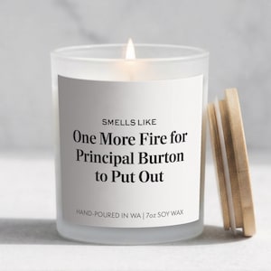 May include: A lit, frosted glass candle with a wooden lid. The candle has a white label with the text "SMELLS LIKE One More Fire for Principal Burton to Put Out." It is hand-poured in WA and contains 7oz of soy wax.