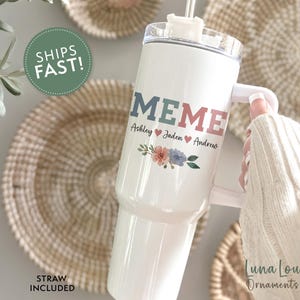 May include: White stainless steel travel mug with a handle and a straw. The mug has the word "MEME" in colorful letters and the names "Ashley, Jaden, Andrew" with hearts between each name. The mug has a floral design with pink, blue, and green flowers.