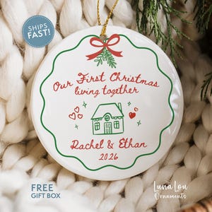 May include: A round white ceramic ornament with a scalloped green border and a gold hanging string. The text reads Our First Christmas living together, Rachel & Ethan, and 2026, accompanied by a green house illustration and red hearts.