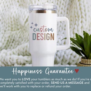 May include: White tumbler with a handle and a clear lid. The tumbler has the words "custom DESIGN" in pastel colors. The tumbler is on a white knitted blanket. A plant in a woven basket is to the right.