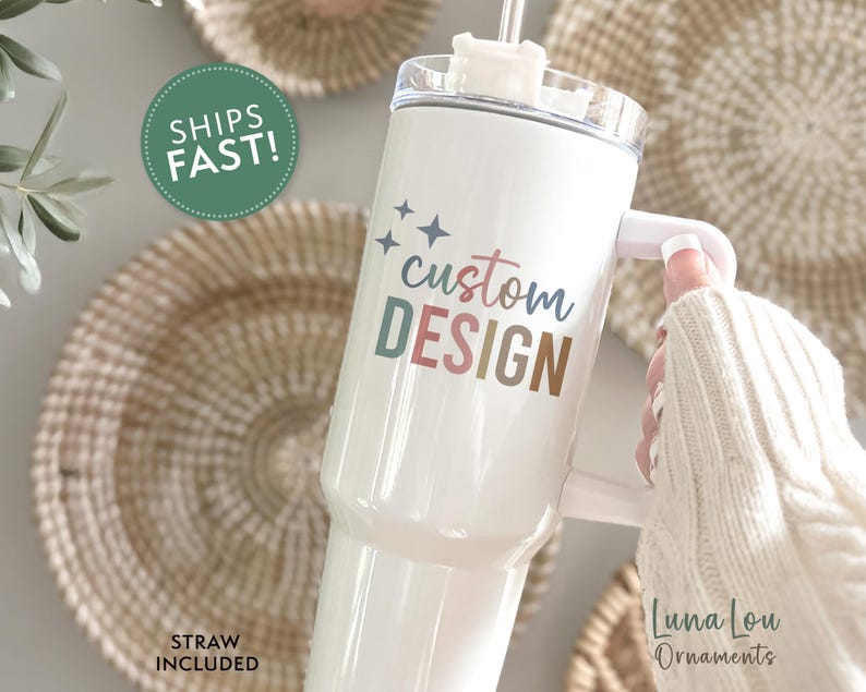 May include: White tumbler with a handle and a straw, featuring the text "custom DESIGN" in pastel colors. The tumbler is held by a person wearing a white sweater. The words "STRAW INCLUDED" are also visible.