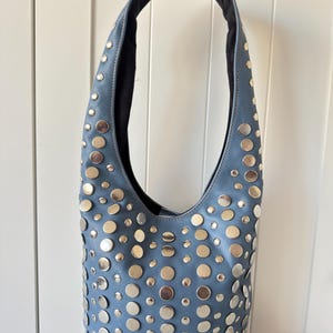 Op de afbeelding: Een blauwe leren schoudertas met zilverkleurige studs van verschillende maten. De tas heeft een gebogen vorm en een enkele schouderriem. De binnenkant is zwart.