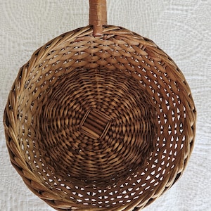 May include: A round, woven wicker basket with a long handle. The basket is a warm brown color and features a circular design with a handle for easy carrying. The basket's open weave allows for ventilation.