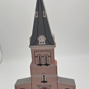 May include: A wooden model of a church with a gray steeple and a clock face. The church is painted in shades of pink and brown with white accents. The sign on the front reads "St. Anne's Church Services 9:00 & 11:00"