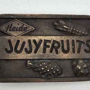 May include: A rectangular, bronze-toned belt buckle with the word "JUJYFRUITS" in large, raised letters. The buckle features the "Heide" logo and embossed fruit designs. The overall design is vintage and detailed.