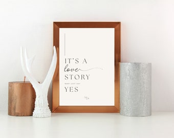 May include: A copper-coloured frame with a white mount and a print that reads "It's a love story. Baby just say YES." The print is on a white background with black text. The frame is sitting on a white surface with a white antler and two candles.
