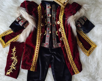 May include: A royal costume set featuring a deep red velvet jacket with gold trim, faux fur accents, and intricate embroidery. The set includes black trousers, a matching vest with red and gold patterns, and a gold crown adorned with faux jewels.